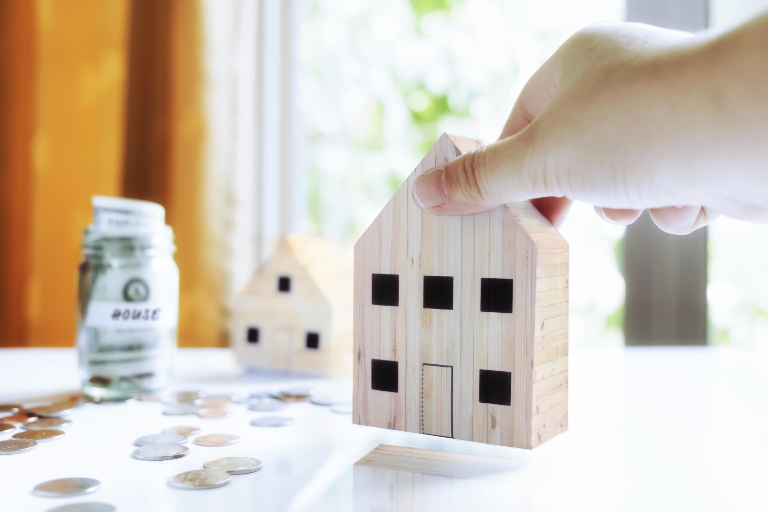 Housing market forecast concept showing a wooden house model, coins, and savings jar representing home prices, affordability, and real estate trends