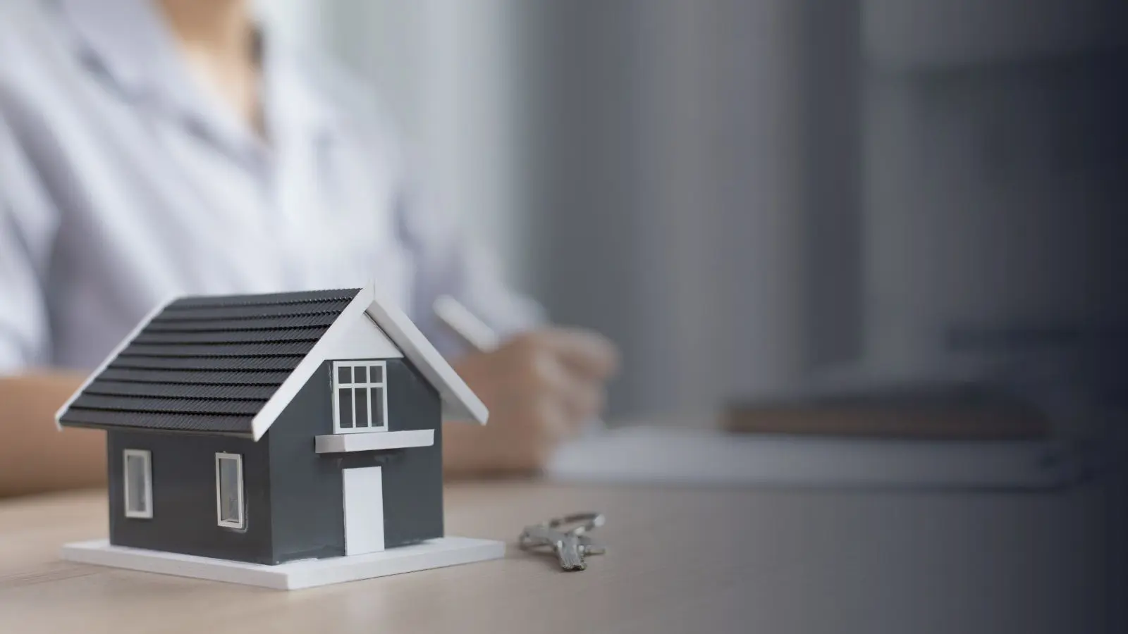 Model house with keys on a desk in front of a person reviewing mortgage paperwork, representing home financing, mortgage lending, and home buying.